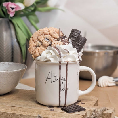 Handgemachte Dessertkerze in einer Porzellantasse mit „Alltagsheld“-Aufschrift, verziert mit Wachssahne, einem Cookie und zwei Schokoriegeln.